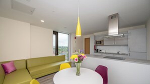 A modern living room with a green sofa, a white dining table with colorful chairs, a yellow pendant light, and a kitchen area with a microwave and stainless steel appliances. - Crow's Nest (Cork)