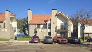 A parking lot with several cars parked in front of a building with red-tiled roofs and a brick facade. - Furnished and Spacious 2-Bedroom Apartment in Downtown Dallas Medical District (Dallas)