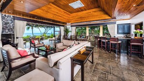 A spacious living area with a wooden ceiling, a stone wall, and a view of the ocean. There are multiple seating areas, a bar, and a television. - Beachfront Villa in  Playa Langosta, Tamarindo! (Playa Langosta)