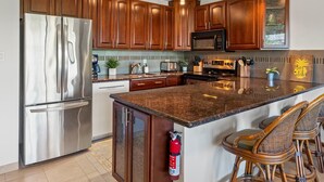 A modern kitchen with wooden cabinets, stainless steel appliances, a large island with bar stools, and a microwave above the counter. - Ocean view condo near Kamaole Beach with pool and lanai summer deals (Kihei)