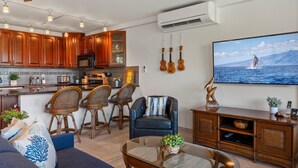A modern living room with a sofa, chairs, a coffee table, and a TV displaying a scenic ocean view. - Ocean view condo near Kamaole Beach with pool and lanai summer deals (Kihei)
