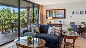 A modern living room with a sofa, coffee table, and chairs. There is a balcony with a view of trees and a ceiling fan. - Ocean view condo near Kamaole Beach with pool and lanai summer deals (Kihei)