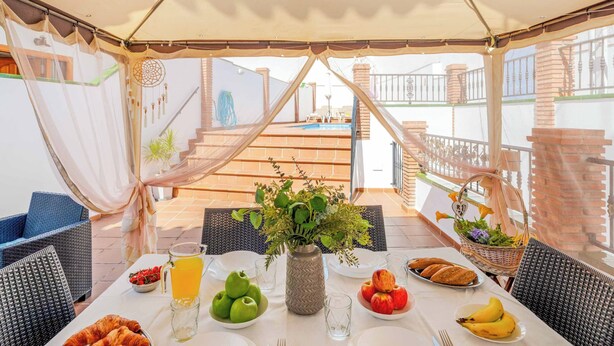 Une table dressée avec des aliments pour le petit-déjeuner, notamment des fruits, des viennoiseries et un vase de fleurs, placée sous une pergola d’où l’on peut admirer une piscine et un escalier.