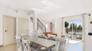 Un vaste espace dédié à la salle à manger, avec une table en verre et des chaises blanches, un escalier et une vue sur le balcon extérieur.