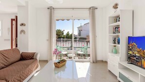 Un salon avec un canapé, une table basse ornée d’une plante, un téléviseur posé sur un meuble et un balcon avec des chaises et une rambarde.