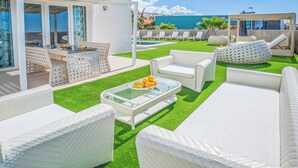 A rooftop patio with white wicker furniture, a glass table with fruit, and a view of the ocean.