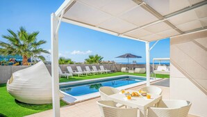 A poolside area with white wicker furniture, a large white bean-shaped sculpture, and a view of the sea.