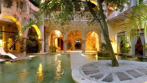 An indoor swimming pool with stone arches, a wooden deck, and a tree in the center. - Casa Pombo 102 (Cartagena)
