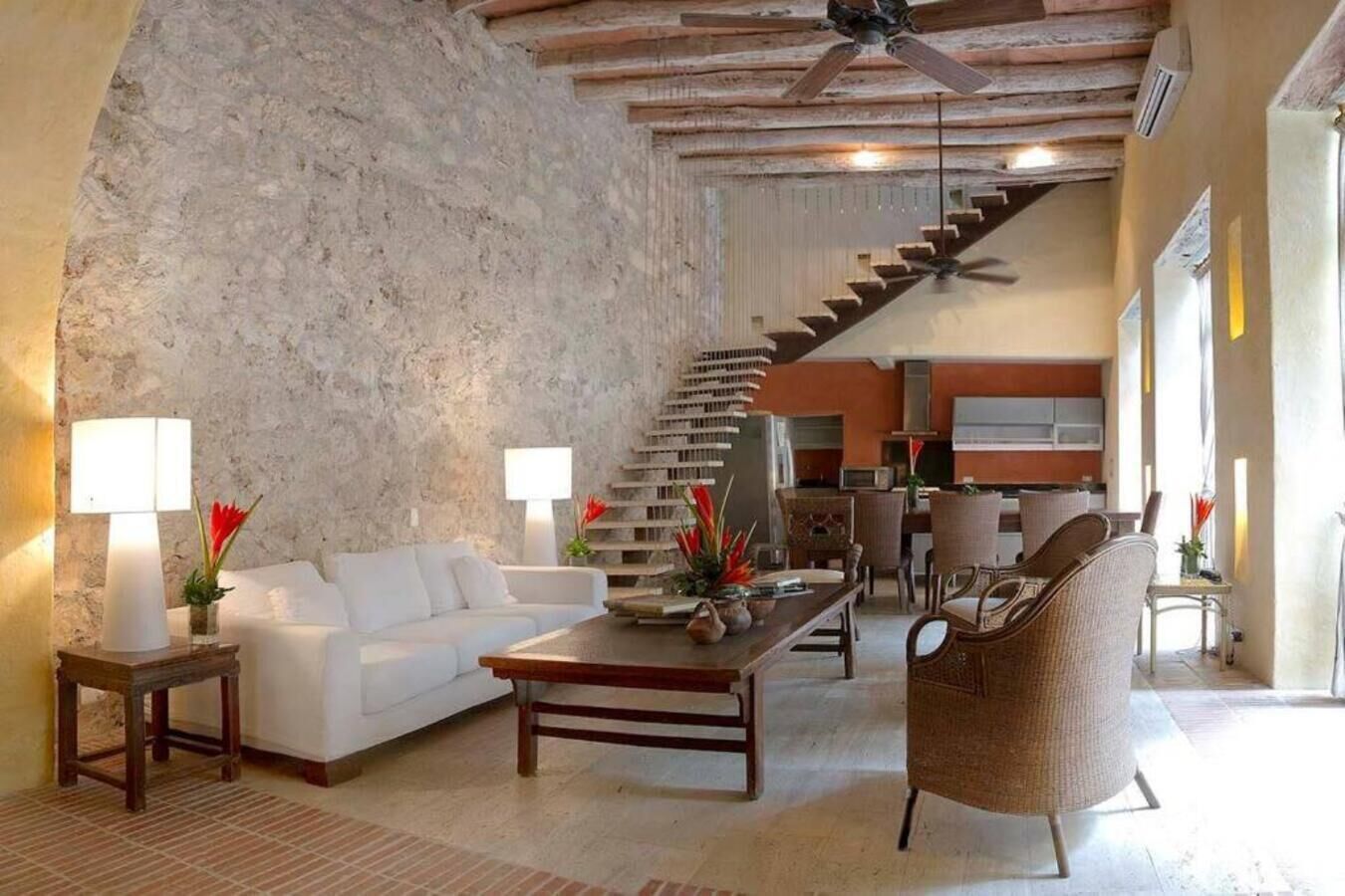 Ein großzügiger Wohnbereich mit einem weißen Sofa, einem Holztisch und Korbstühlen. Eine Treppe führt zu einer oberen Ebene.