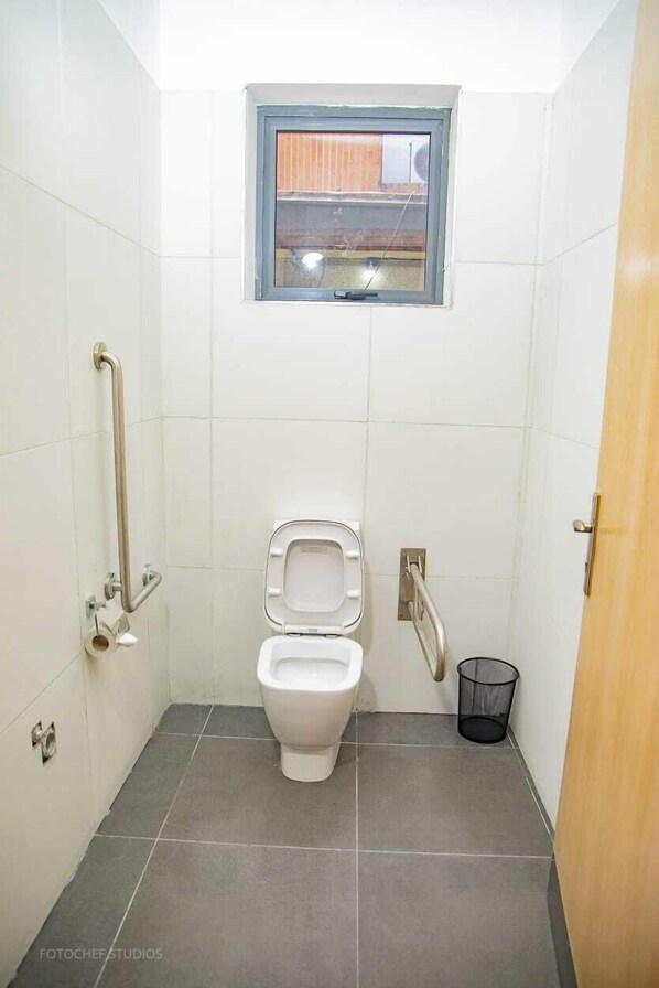 A small, tiled bathroom with a toilet, grab bars, a window, and a trash can. - Jersey Apartments By Genesis  (Port Harcourt)