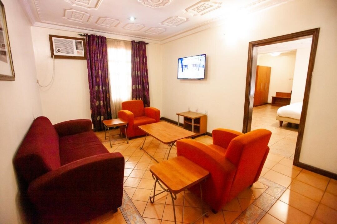 A hotel room with a red sofa, two orange armchairs, a wooden coffee table, and a TV mounted on the wall.