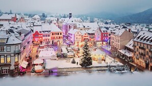 A snow-covered town square with historic buildings, a prominent Christmas tree, and a river in the foreground.