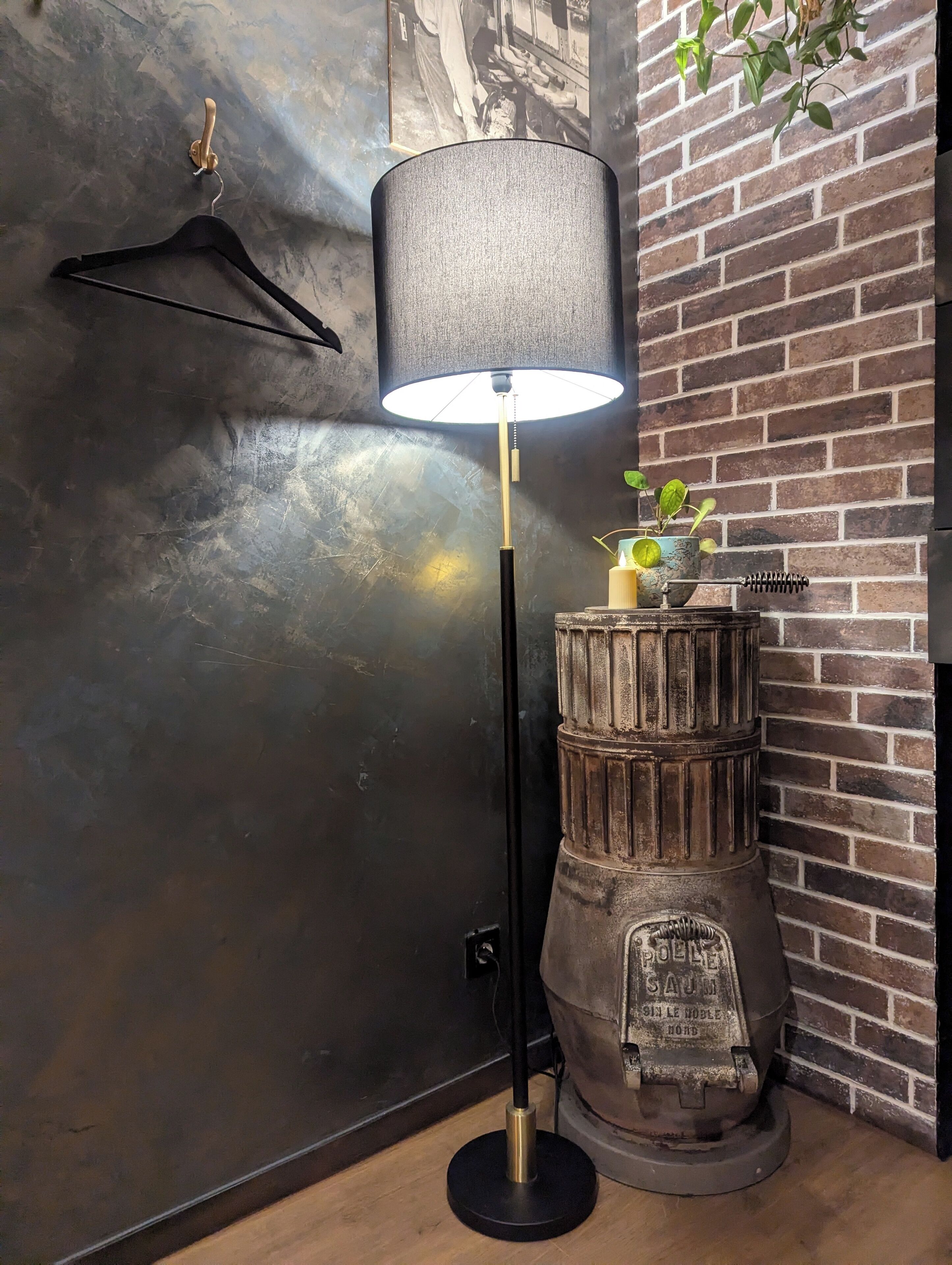 A lamp with a black base and a white shade, a wooden barrel with a candle and a plant on top, and a brick wall.