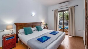 Ein Hotelzimmer mit Bett, Nachttisch, Lampe, Wand-Klimaanlage und Blick auf einen Außenbereich mit Pool und Palmen.