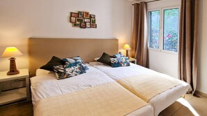 A double bed with a quilted cover, two bedside tables with lamps, a wall-mounted picture frame, and a window with curtains. - Villa Casa Amelia by Villa Plus (Canico)