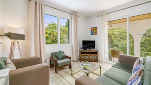 A modern living room with a sofa, armchair, and a glass coffee table. There is a television on a wooden cabinet, and large windows offer a view of greenery outside. - Villa Casa Amelia by Villa Plus (Canico)