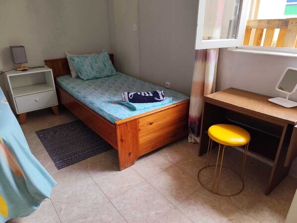 A small, single-bed room with a wooden bed, a desk with a chair, a nightstand, and a window with a view.