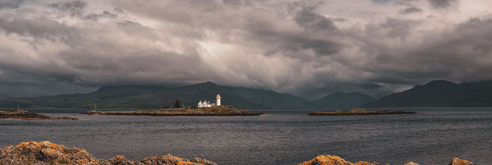 A coastal scene with a lighthouse on a small island, surrounded by mountains and a cloudy sky.