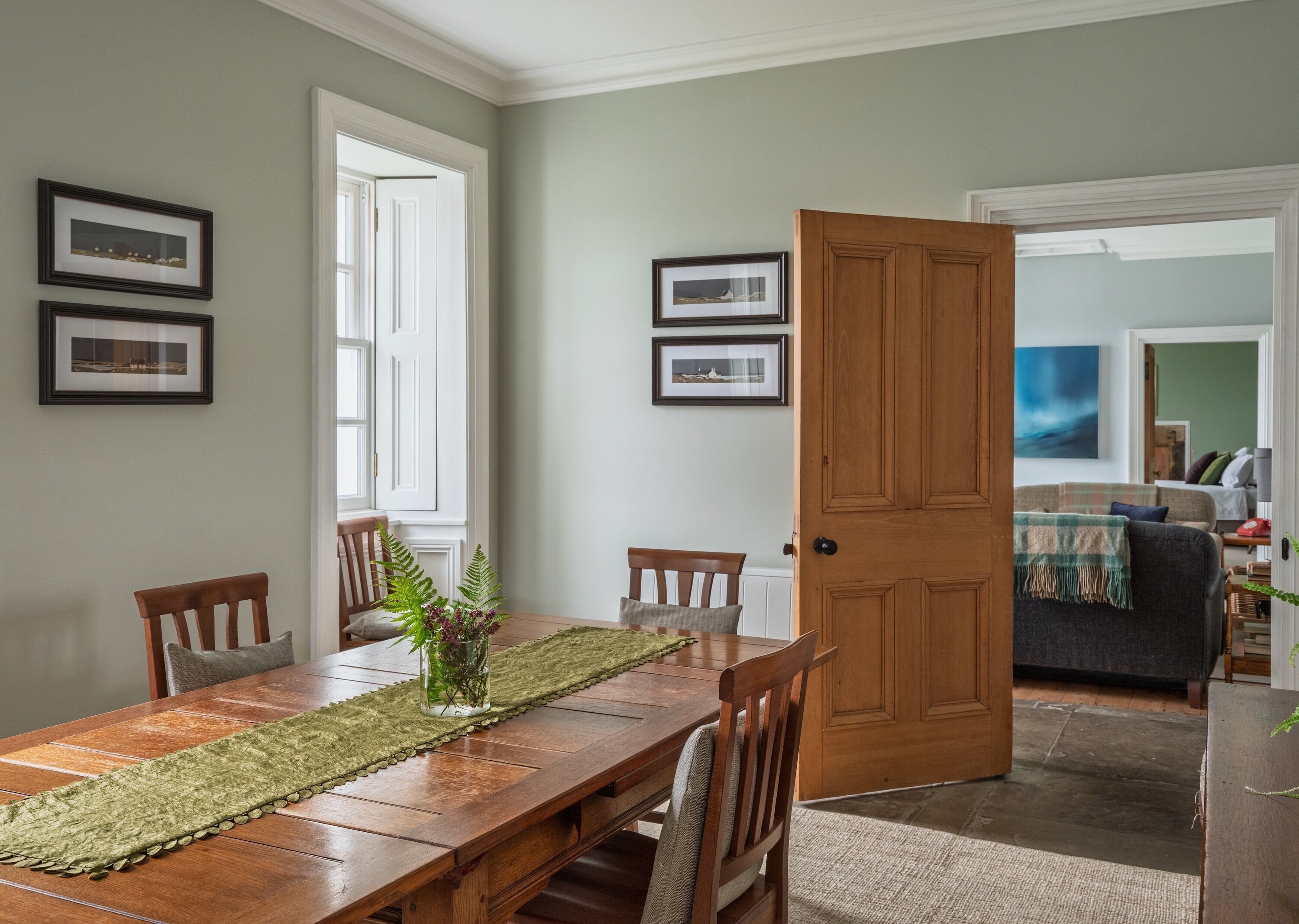A dining area with a wooden table, chairs, and a green runner. There are framed pictures on the walls and a door leading to another room.