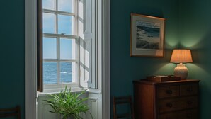 Een slaapkamer met een groot raam, een nachtkastje met een plant, een kaptafel en een lamp.