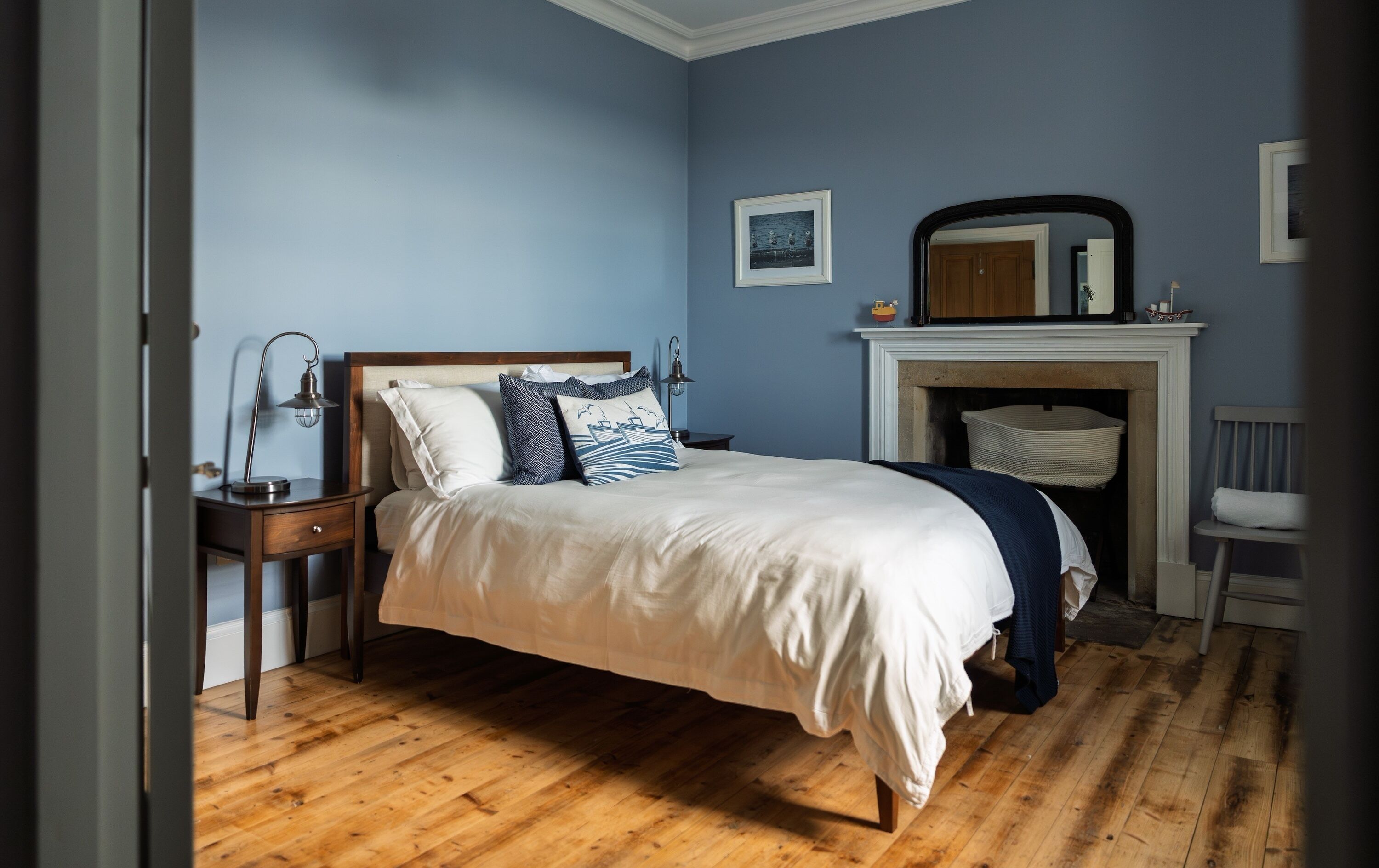 Una camera da letto con pavimento in legno, un letto con lenzuola bianche e azzurre, un comodino con una lampada, un camino e uno specchio appeso al muro.