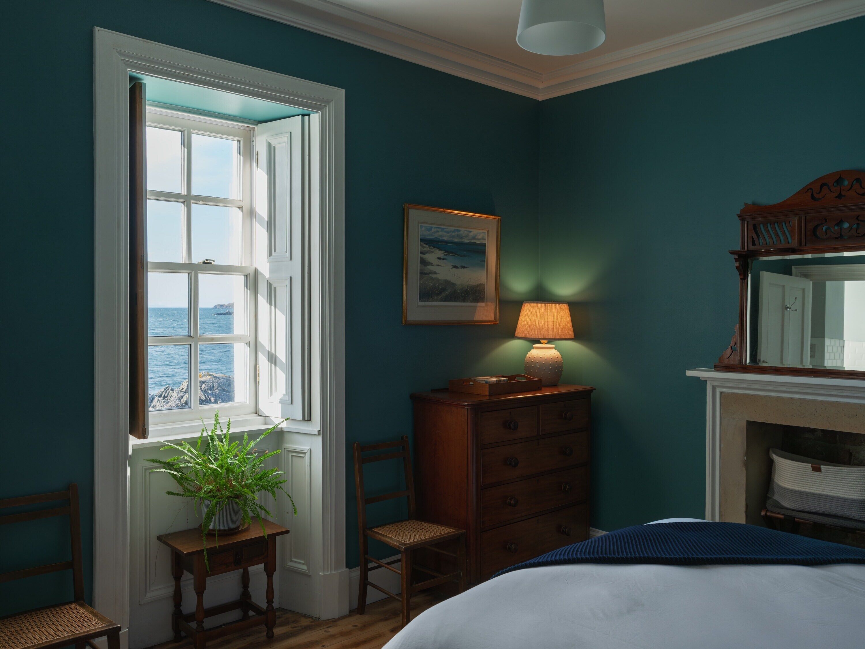 A bedroom with a large window, a dresser, a chair, a bedside table with a lamp, and a mirror.