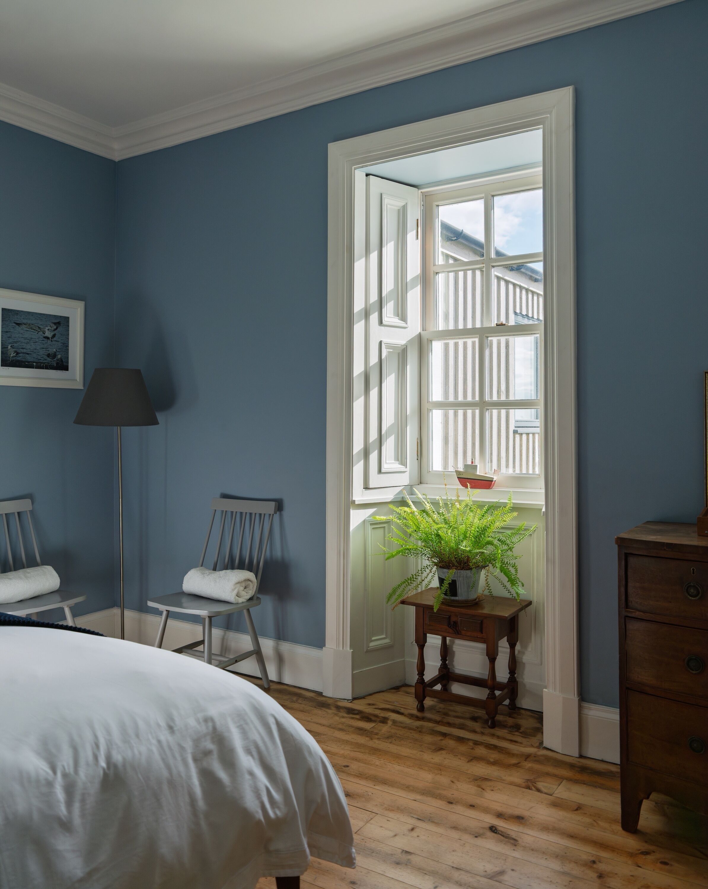 A bedroom with a large window, a bed, a chair, a lamp, and a dresser.