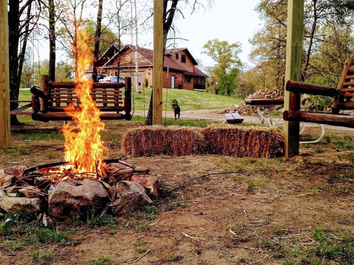A campfire with hay bales and a wooden bench in a wooded area with a house in the background.
