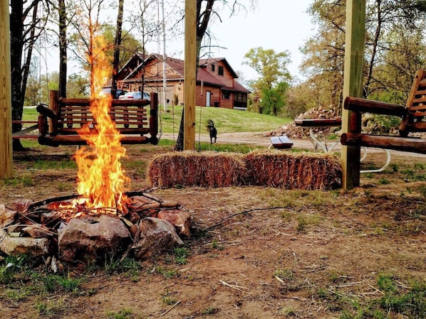A campfire with hay bales and a wooden bench in a wooded area with a house in the background.