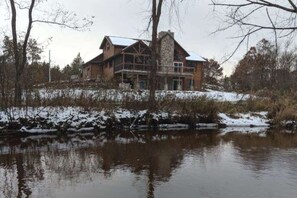 A large, multi-story log cabin with a stone chimney, situated by a calm river with snow on the banks.