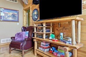 Une pièce chaleureuse avec un fauteuil en cuir, des étagères en bois remplies de livres et de jouets, un tableau accroché au mur et un téléviseur à écran plat.