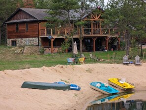 A cabin with a stone foundation, wooden exterior, and a deck. There are chairs and a table on the deck, and a boat on the sand near the water. - The Land in the Pines-Cabin (Necedah)