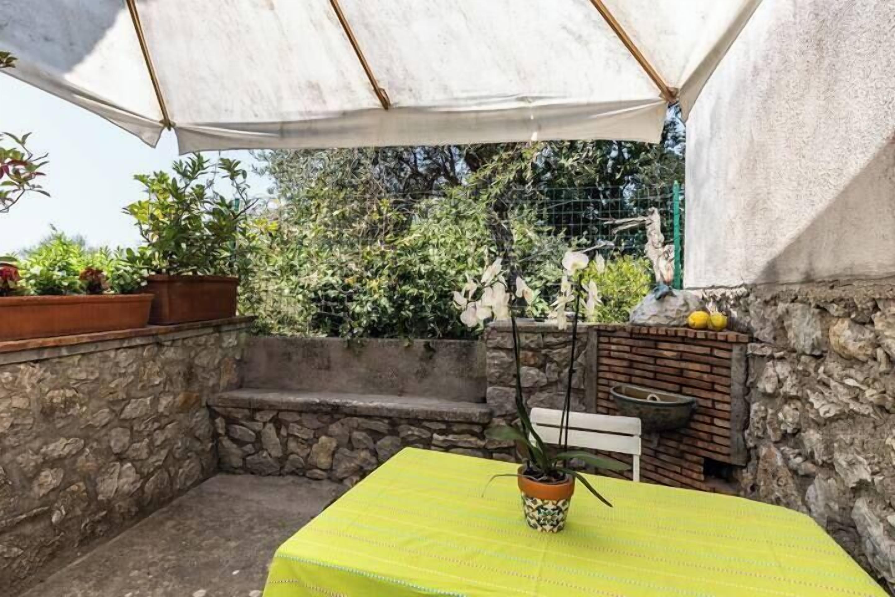 Un pequeño patio con una mesa cubierta por un paño amarillo, una planta en maceta y un muro de piedra.