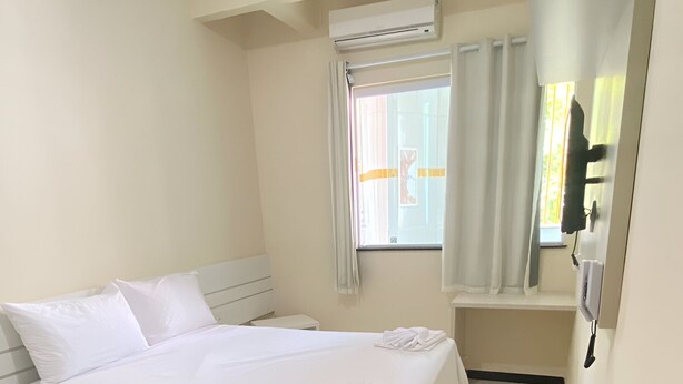 A hotel room with a bed, a small table, a mirror, and a window with curtains. - Palladium Pousada (Neopolis)