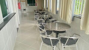 A corridor with a row of round tables and chairs, a fire extinguisher, and a window with curtains. - Palladium Pousada (Neopolis)