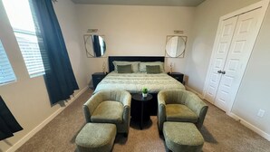 Un dormitorio con una cama, dos mesitas de noche, dos sillas, una ventana con cortinas y una puerta.