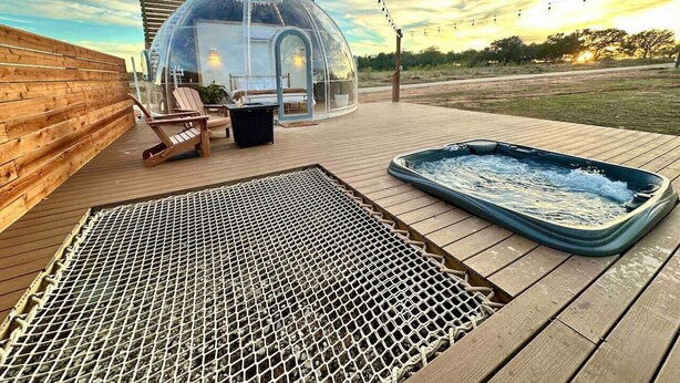 Een transparante kasconstructie met een hottub en zitgedeelte op een houten terras.