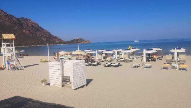 Ein Strand mit Liegestühlen und Sonnenschirmen, einem Rettungsturm und freiem Blick auf das Meer und ferne Berge.