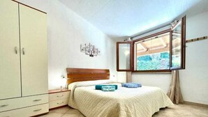 Ein Schlafzimmer mit Bett, Kleiderschrank, Nachttisch, Fenster mit Blick und Deckenleuchte.