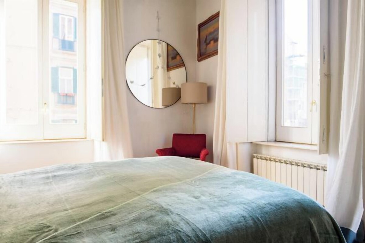A room with a bed, a red chair, a round mirror, and a window with curtains.