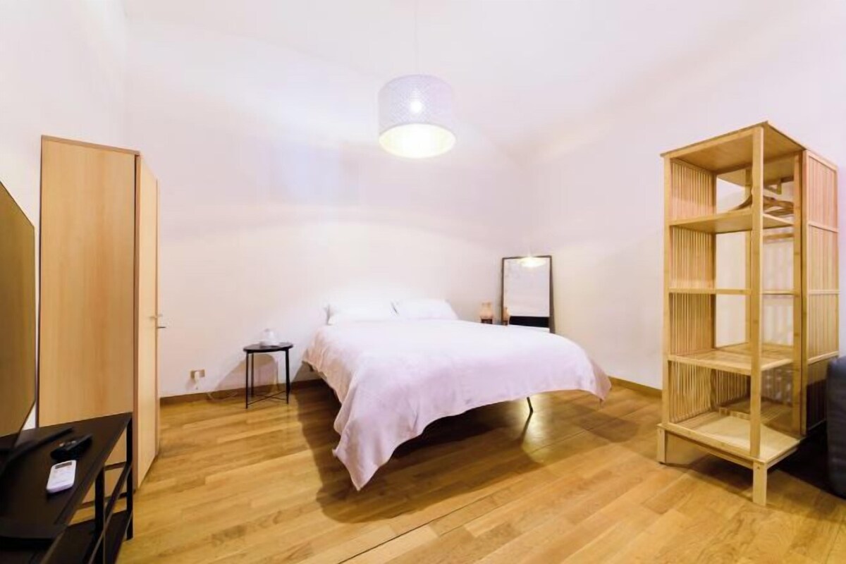 A bedroom with a bed, a wooden wardrobe, a small table, a bookshelf, and a hanging light fixture.
