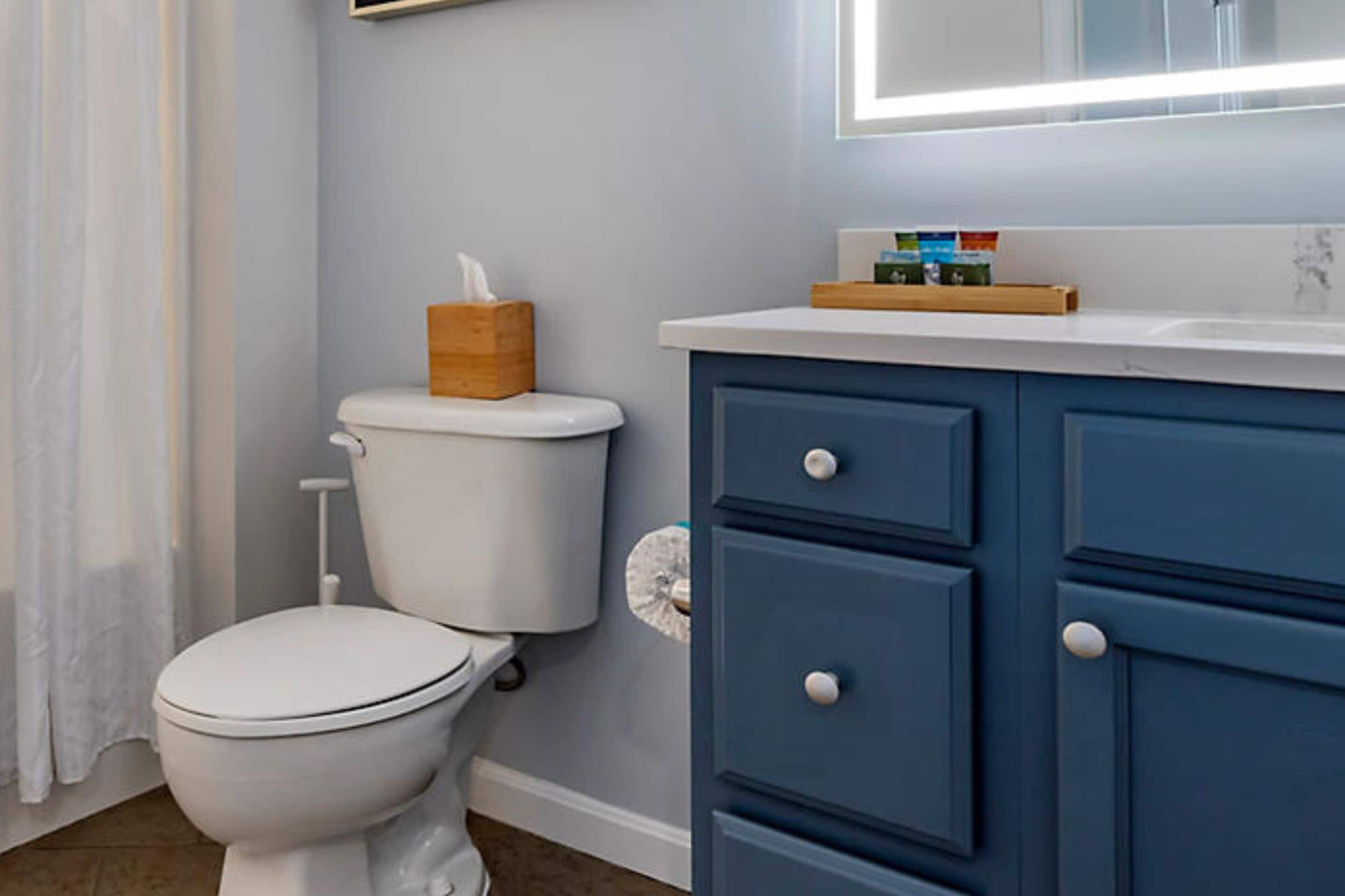 Un baño con inodoro blanco, un mueble azul con cajones y un espejo sobre el lavamanos.