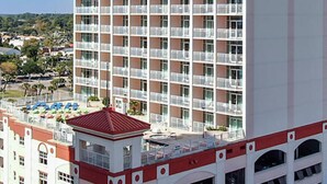 A multi-story hotel with balconies, a rooftop pool area, and a red-roofed structure. - Carolina Grande (Myrtle Beach)