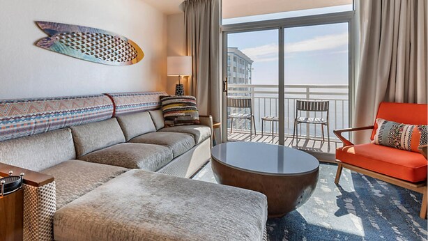 A hotel room with a sofa, a round coffee table, a red armchair, a balcony with chairs, and a decorative wall piece. - Carolina Grande (Myrtle Beach)