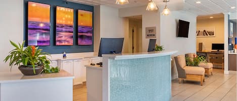 A reception area with a curved counter, a large wall with three sunset paintings, and a seating area with two chairs and a plant.