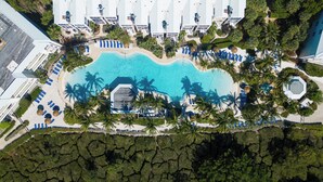 An aerial view of a resort with a central swimming pool surrounded by palm trees and sun loungers.