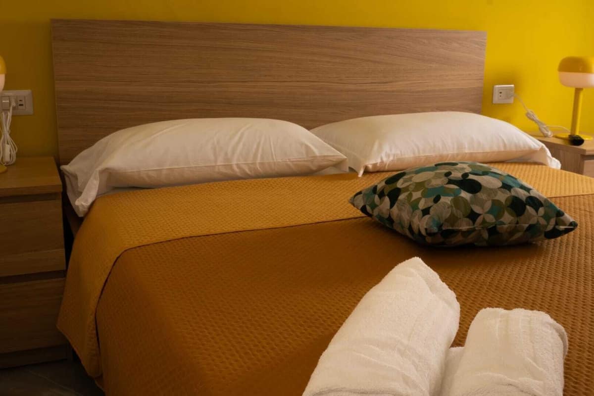 A neatly made bed with a brown bedspread, two white pillows, and a camouflage pillow. There are two bedside tables with lamps and a phone.