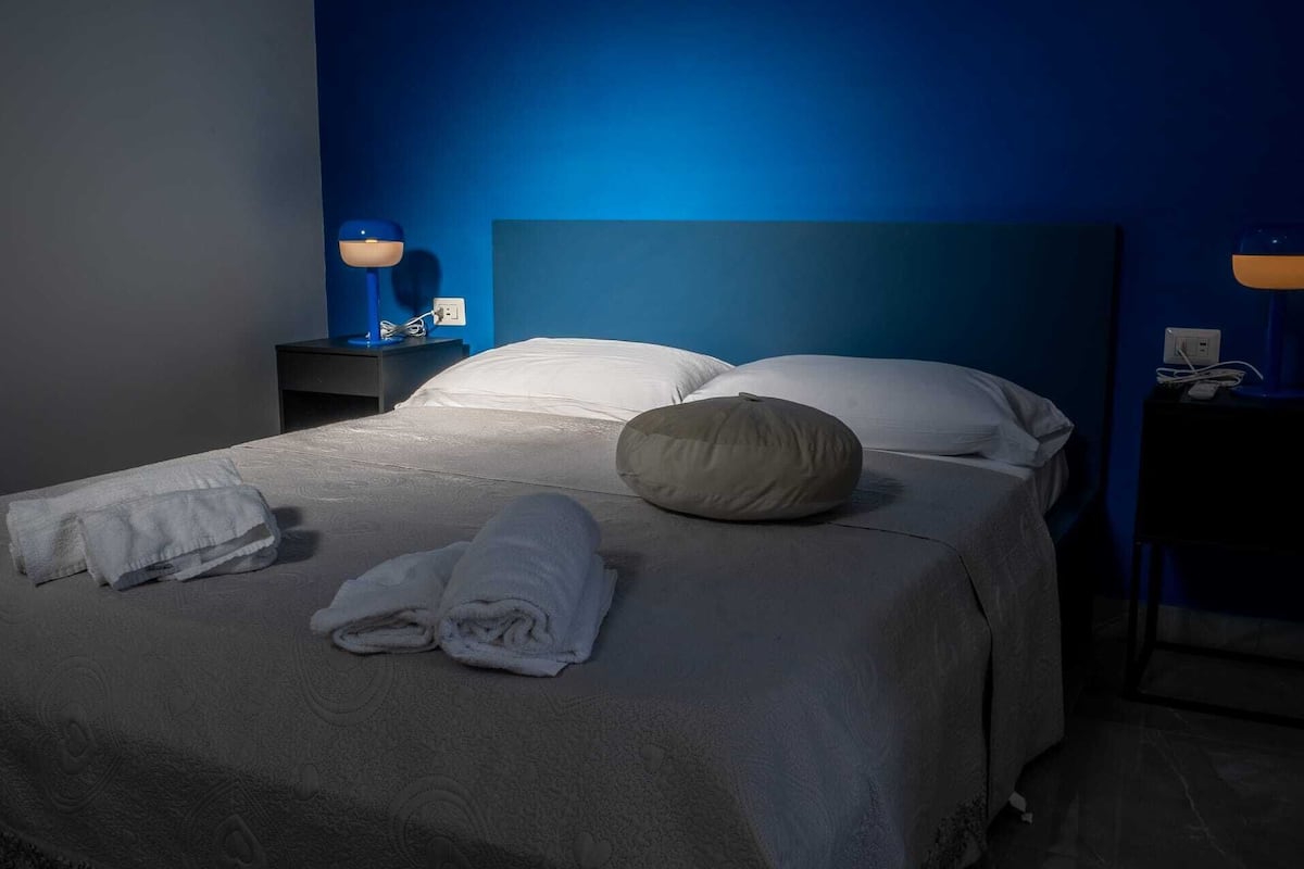 A neatly made bed with white linens and a beige pillow, a blue wall, and a nightstand with a lamp.