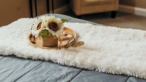 Una camera da letto con un letto, un tappeto bianco e soffice, un vassoio di legno con girasoli, un tavolino con un vaso e una sedia.