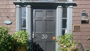 Een zwarte deur met het nummer 30, geflankeerd door twee grote plantenbakken met bloemen, die leidt naar een houten trap.